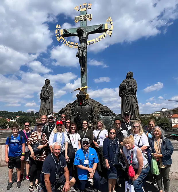 Tekton pilgrims in front of the Statue of the Crucifixion and Calvary on Charles Bridge in Prague, with Christ flanked by Mary and St. John the Evangelist beneath the gold Hebrew Sanctus inscription.