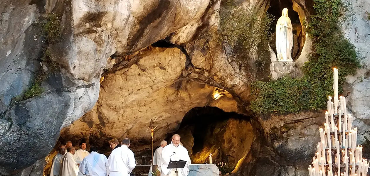 Priest preaching at Mass before the Grotto of Massabielle in Lourdes, concelebrants at the altar beneath the statue of Our Lady of Lourdes