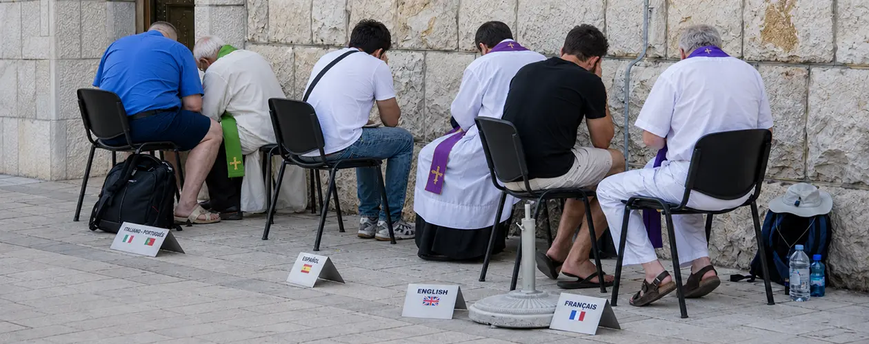 Catholic priests in white albs and purple stoles hearing outdoor confessions along a stone church wall, with English, Spanish, French and Italian language signs for pilgrims