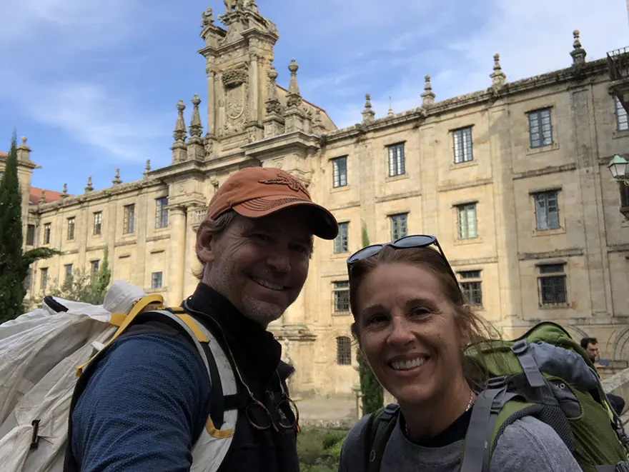 Krista Behringer and her husband wearing backpacks in front of a stone monastery facade in Santiago de Compostela at the end of their Camino de Santiago.